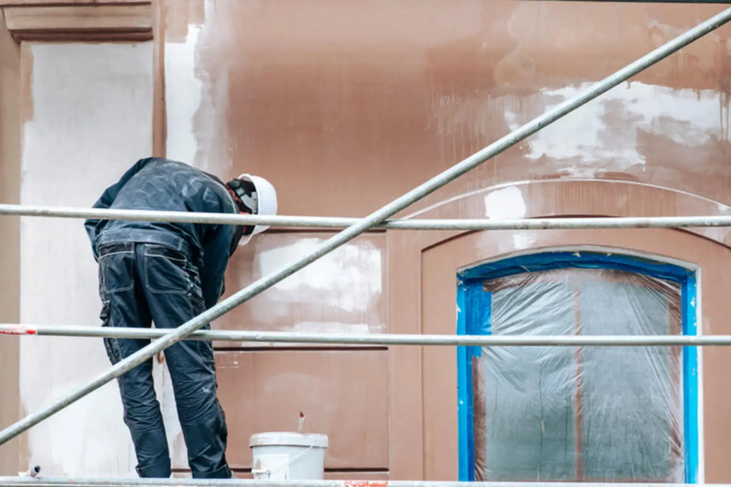 Painter on scaffolding preparing a masked masonry building facade for a fresh coat, showing how often a commercial building should be repainted.