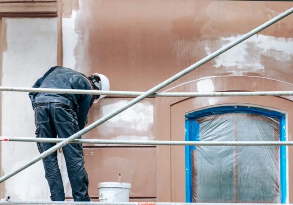 Painter on scaffolding preparing a masked masonry building facade for a fresh coat, showing how often a commercial building should be repainted.
